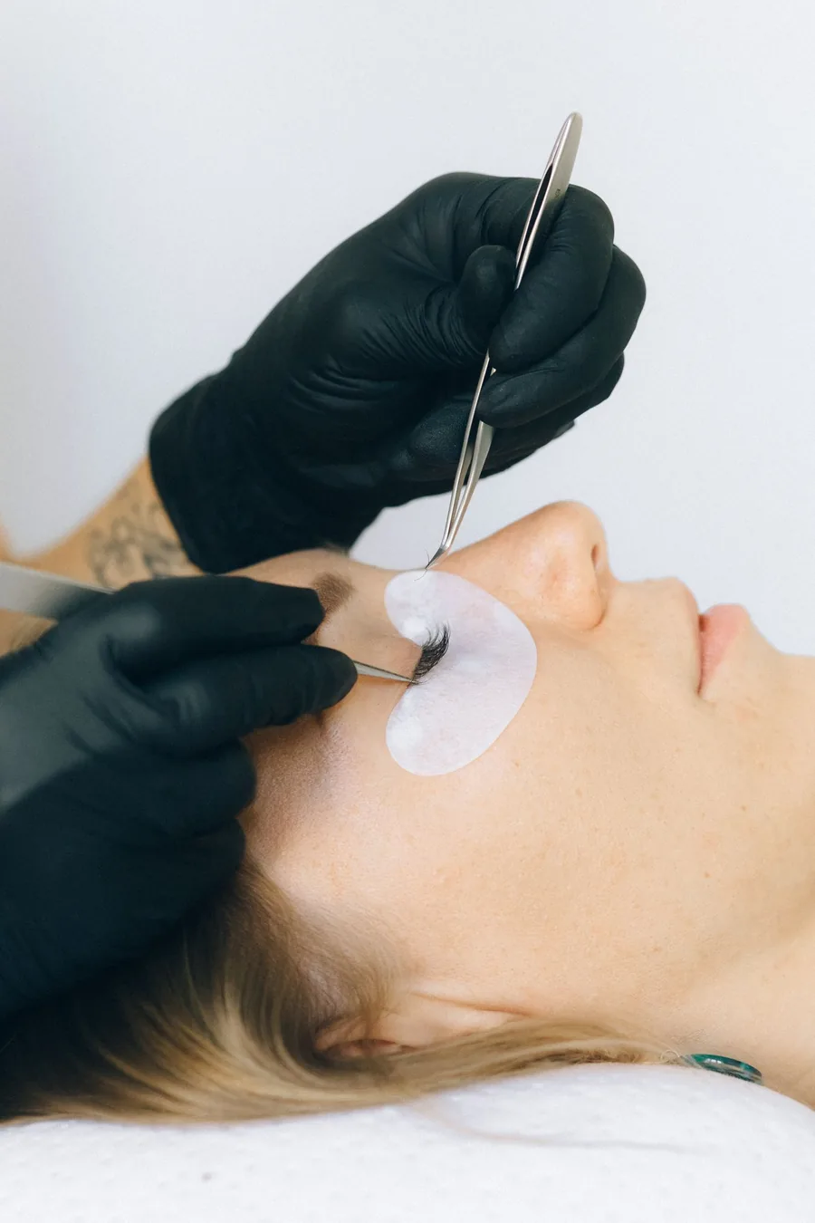 Lash technician in black gloves applying extensions to a reclining client against a clean white background